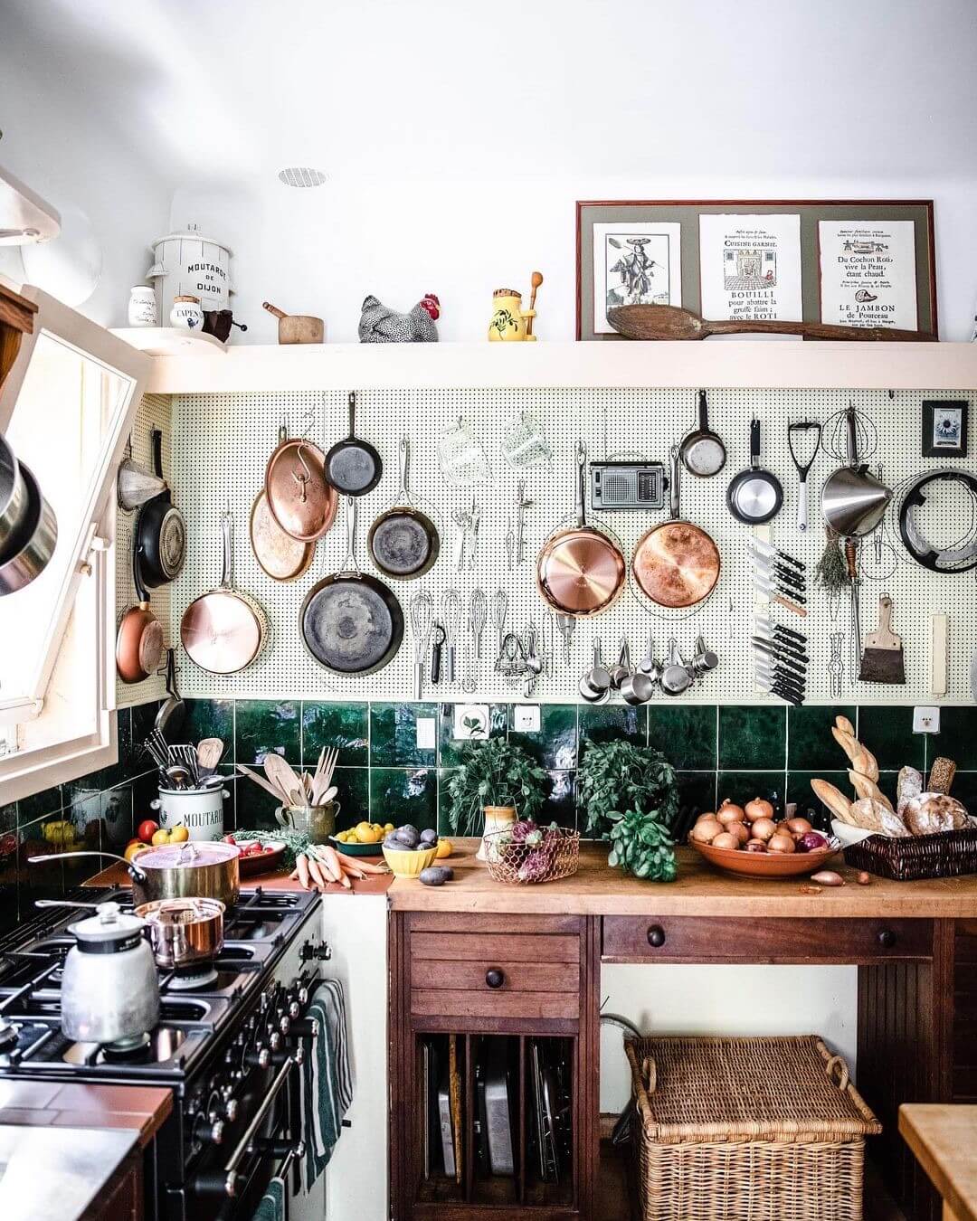 Quaint kitchen in french home, where cooking classes are offered. There are copper pots and iron skillets hanging on the walls above a green tiled backsplash and rustic wooden countertops.